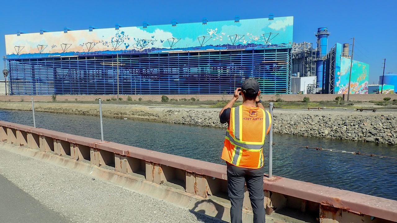 Worker wearing a hat and safety vest taking a picture of a water desalination plant along a channel